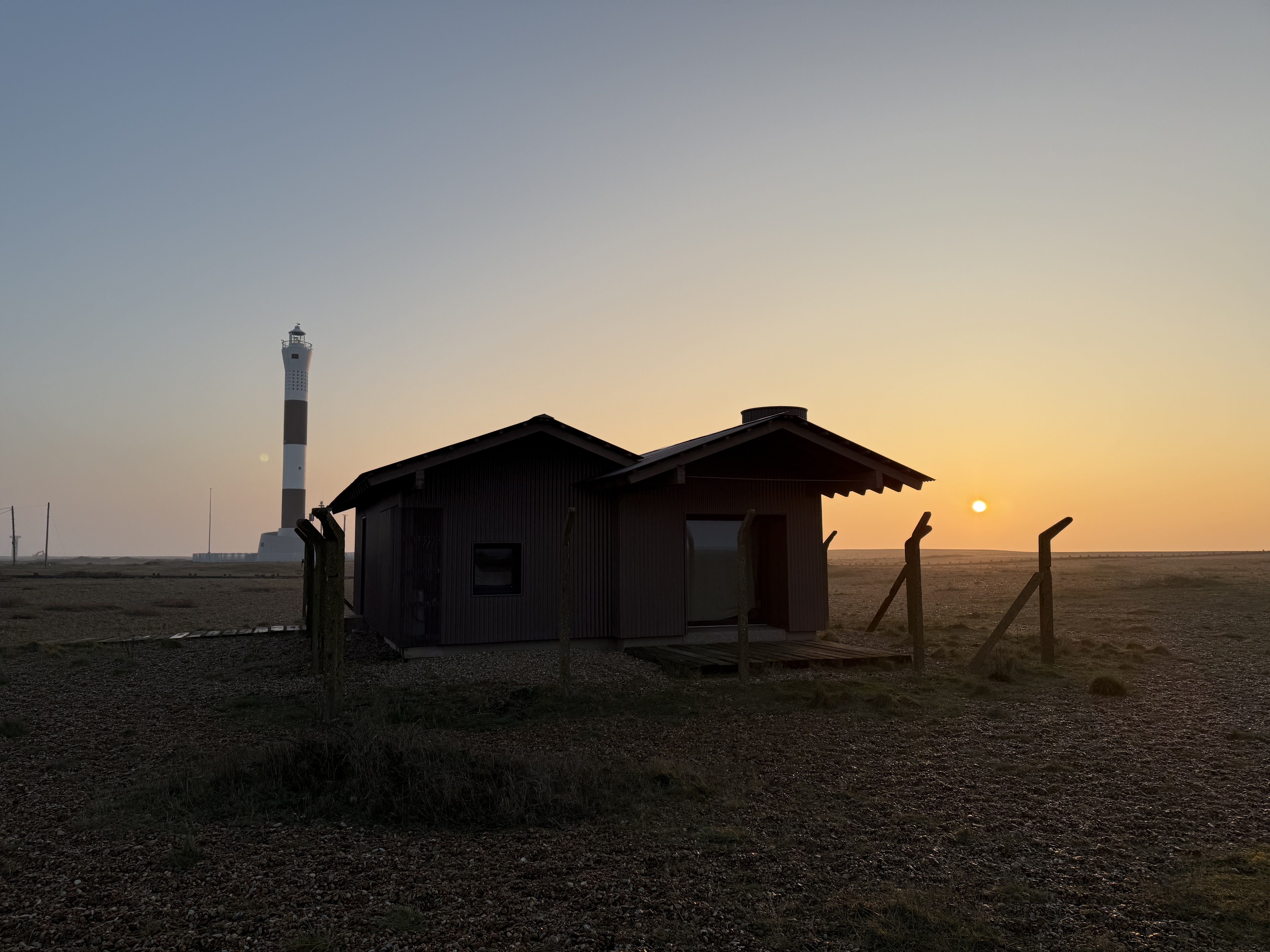 An image of 'Radar' a converted radar station on Dungeness beach that is now a holiday home. It is red rust coloured corrugated building, with large picture windows. It is in the sunrise over the beach with a black and white lighthouse in the background.