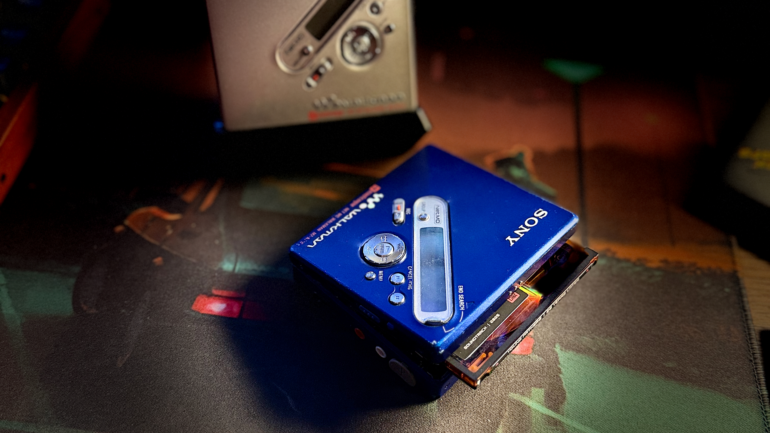 An image of two Sony MiniDisc players sat on a desk, one silver in colour is upright in a stand. The other a deep blue colour is laying on the desk, open with a disc poking out.